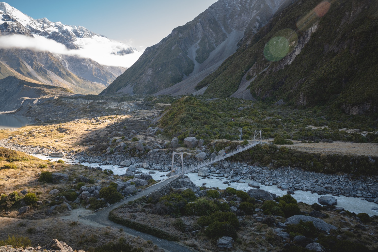The Hooker Valley: a swing bridge over a pale glacial river, boulders and scrub, steep mountainsides, and snowy peaks under a band of cloud