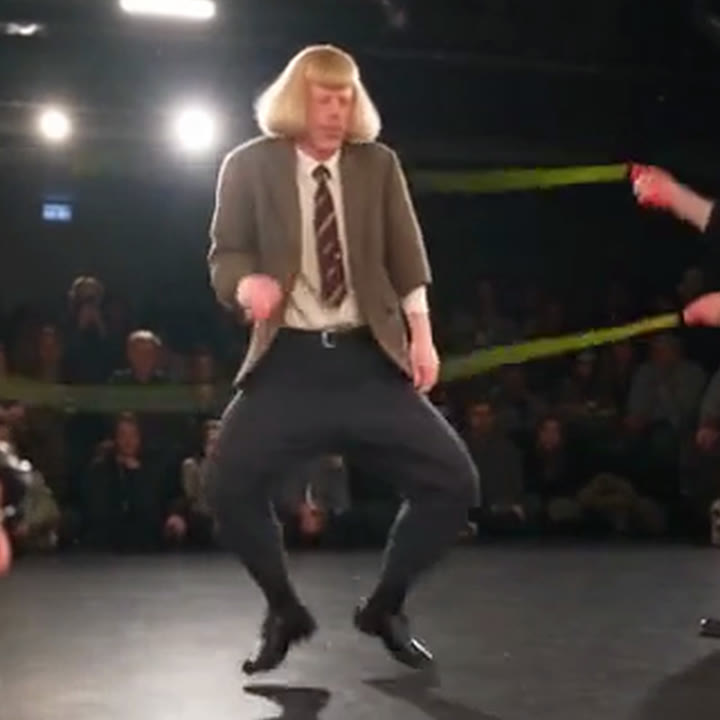 The original performer in a blond wig and tweed jacket, mid-hop in a bow-legged crouch between the ropes